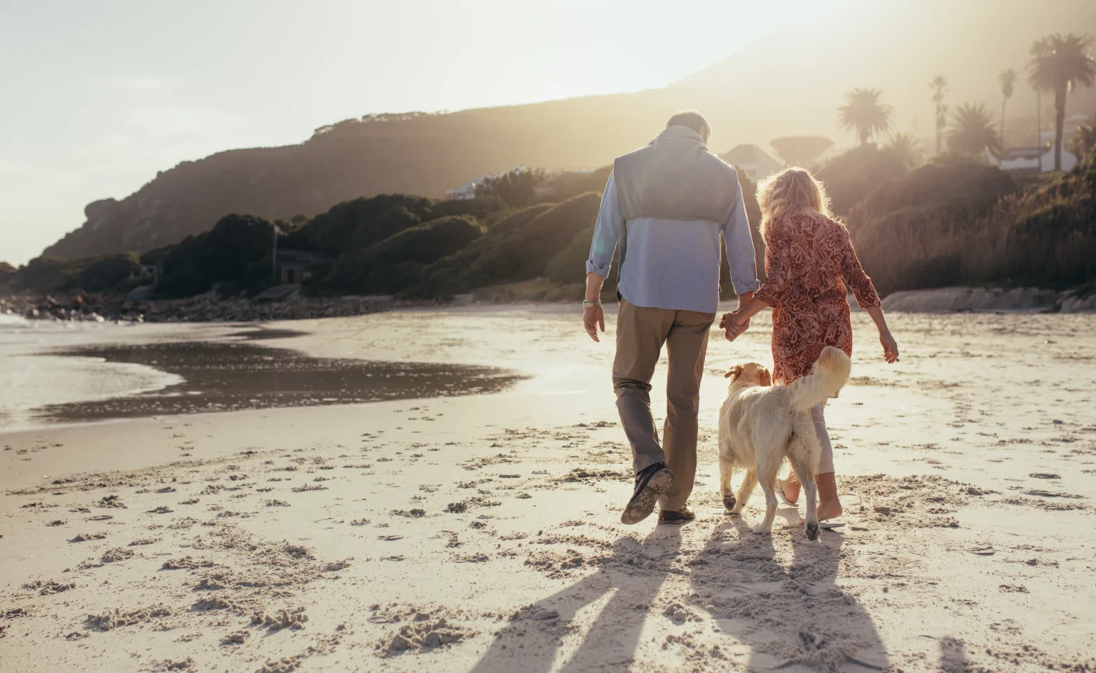 A man and woman hold hands on the beach with their golden retriever following them